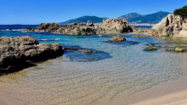 Une plage avec une eau claire et peu profonde, des formations rocheuses et une chaîne de montagnes visible au loin.