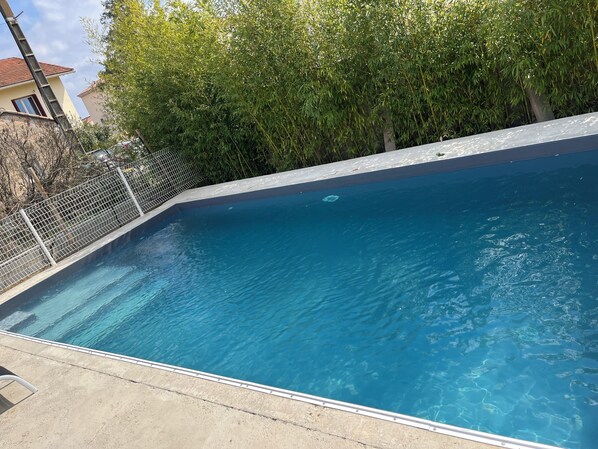 A swimming pool with a clear blue water surface, surrounded by a metal fence and lush green trees.
