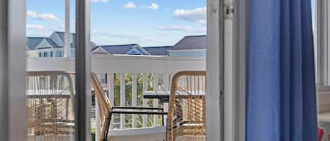 Un balcon avec des fauteuils en osier et une table, donnant sur un quartier résidentiel avec des maisons et un ciel dégagé.