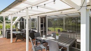 Un patio techado al aire libre con mesa y sillas, un sofá y un ventanal de vidrio que da a un jardín.