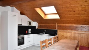 Een kleine keuken met witte kasten, een magnetron, een fornuis en een spoelbak. Er staat een houten eettafel met stoelen en een rode tafelkleed. Een dakraam zorgt voor natuurlijk licht.