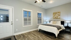 Un dormitorio con una cama, ventilador de techo, dos ventanas con persianas, una puerta y una obra de arte colgada en la pared.