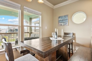 Un coin repas comprenant une grande table en bois, deux chaises et un banc. Il y a deux fenêtres équipées de stores, un tableau accroché au mur et un miroir rond.