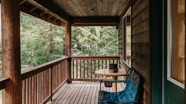 A wooden porch with a table and a blue chair, overlooking a forested area. - 77mbr - Wi-fi - Fireplace - Pets Ok - Bbq 3 Bedroom Cabin by RedAwning (Deming)