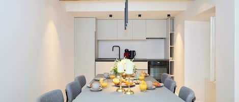 A modern dining area with a long table, grey chairs, and a kitchen in the background.