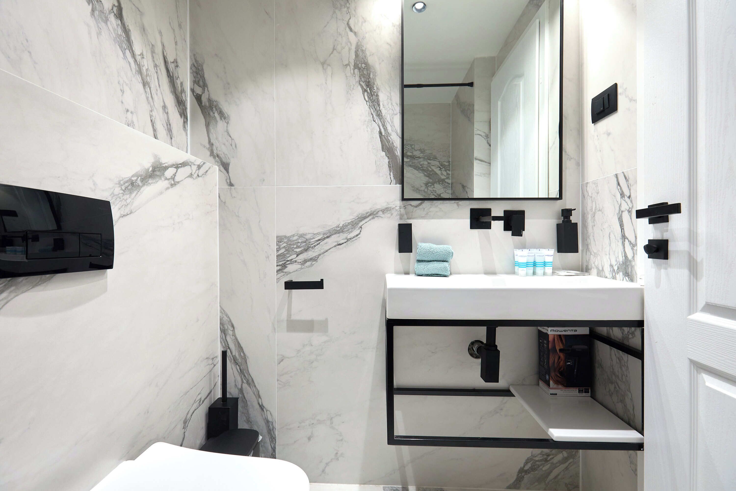 Une salle de bain moderne avec des murs en marbre, un lavabo blanc, des accessoires noirs et un miroir.