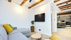 A modern living room with a grey sofa, wooden beams on the ceiling, a flat-screen TV, and a small dining area in the background. - Black Water Bay house (Dubrovnik)