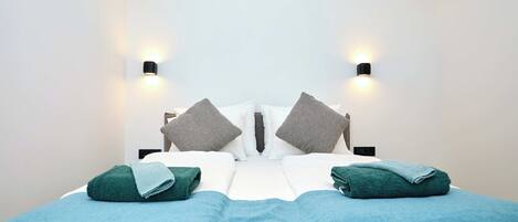 A neatly made bed with two pillows, a blue blanket, and two folded towels. There are two wall-mounted light fixtures on either side of the bed.