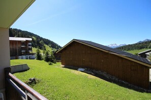 A wooden building with a sloped roof, surrounded by a grassy area and a mountain backdrop.