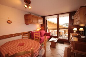 A cozy room with a wooden table and chairs, a small sofa, a balcony with a view, and wooden cabinets.