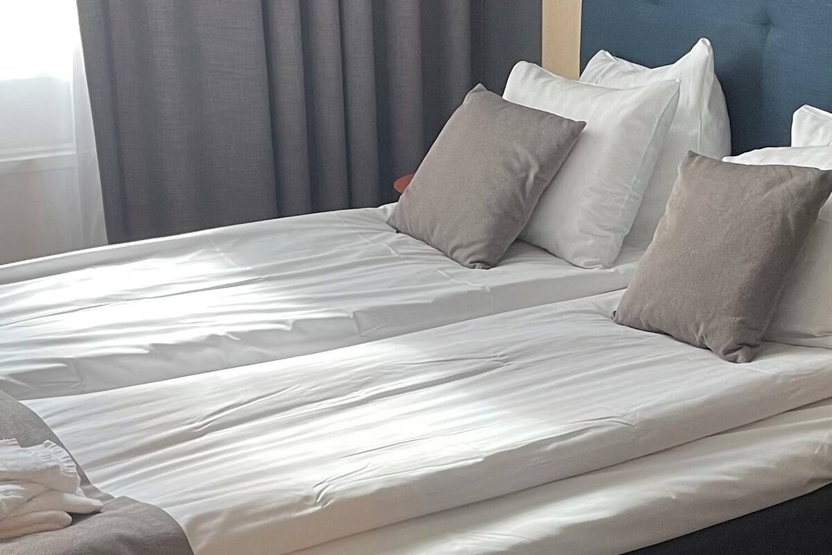 A neatly made bed with white linens and two pillows, a dark headboard, and grey curtains in a hotel room.
