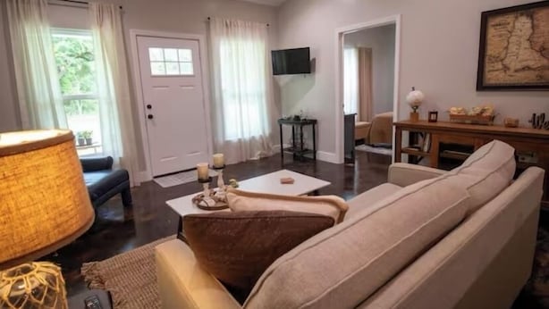 A living room with a beige sofa, a coffee table, and a lamp. There is a TV mounted on the wall and a door leading to another room. - Hundred Mullet Hotel- New Home Near Foley  (Foley)