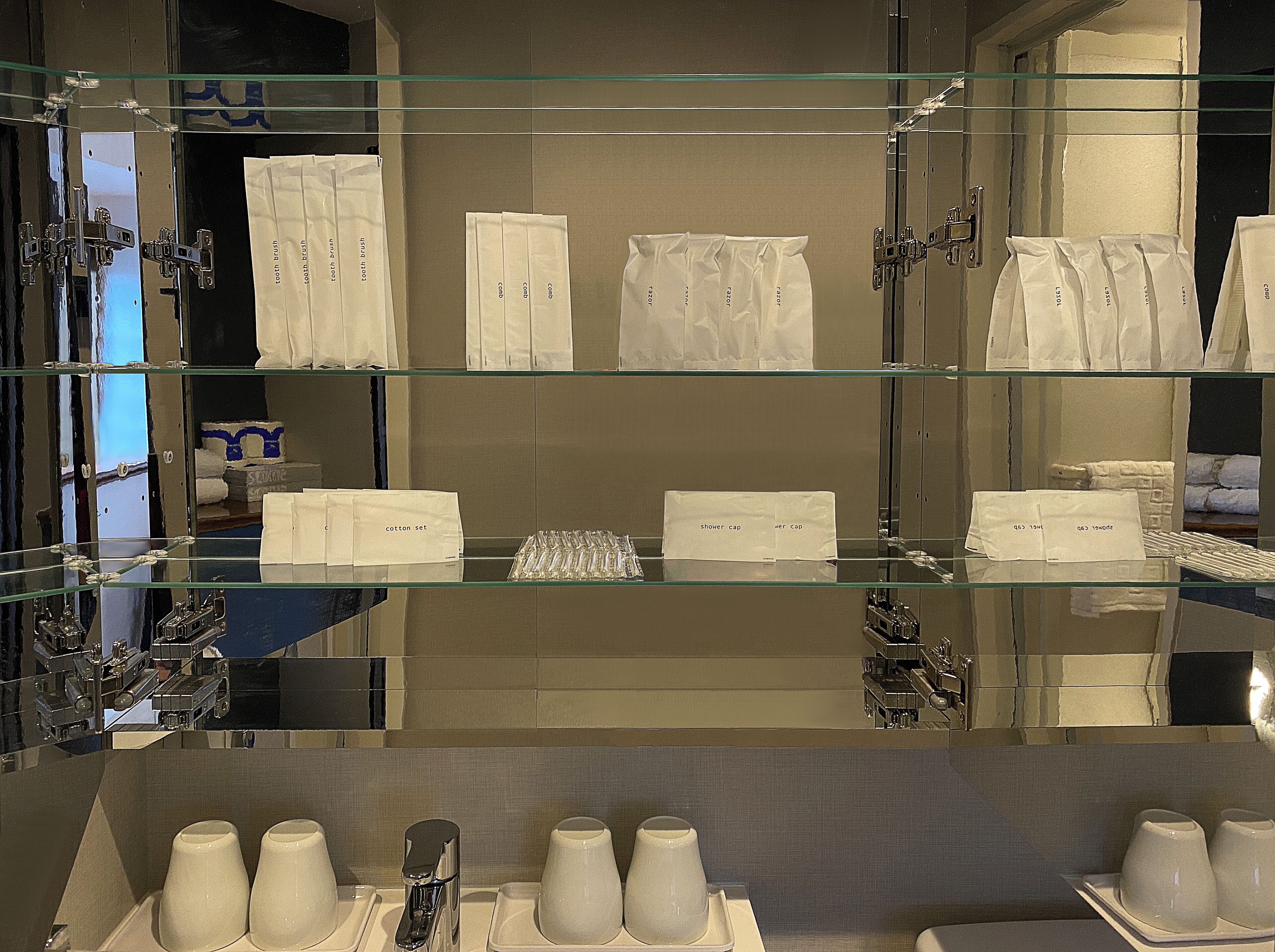 A hotel bathroom with glass shelves displaying toiletries like toothbrushes, cotton sets, and shower caps.
