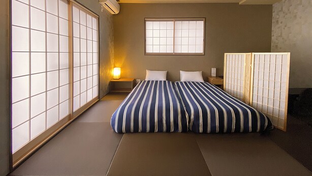 A traditional Japanese-style bedroom with tatami flooring, a shoji screen, a bed with striped bedding, and a bedside table with a lamp. - My Home Inn Izumisano (Izumisano)