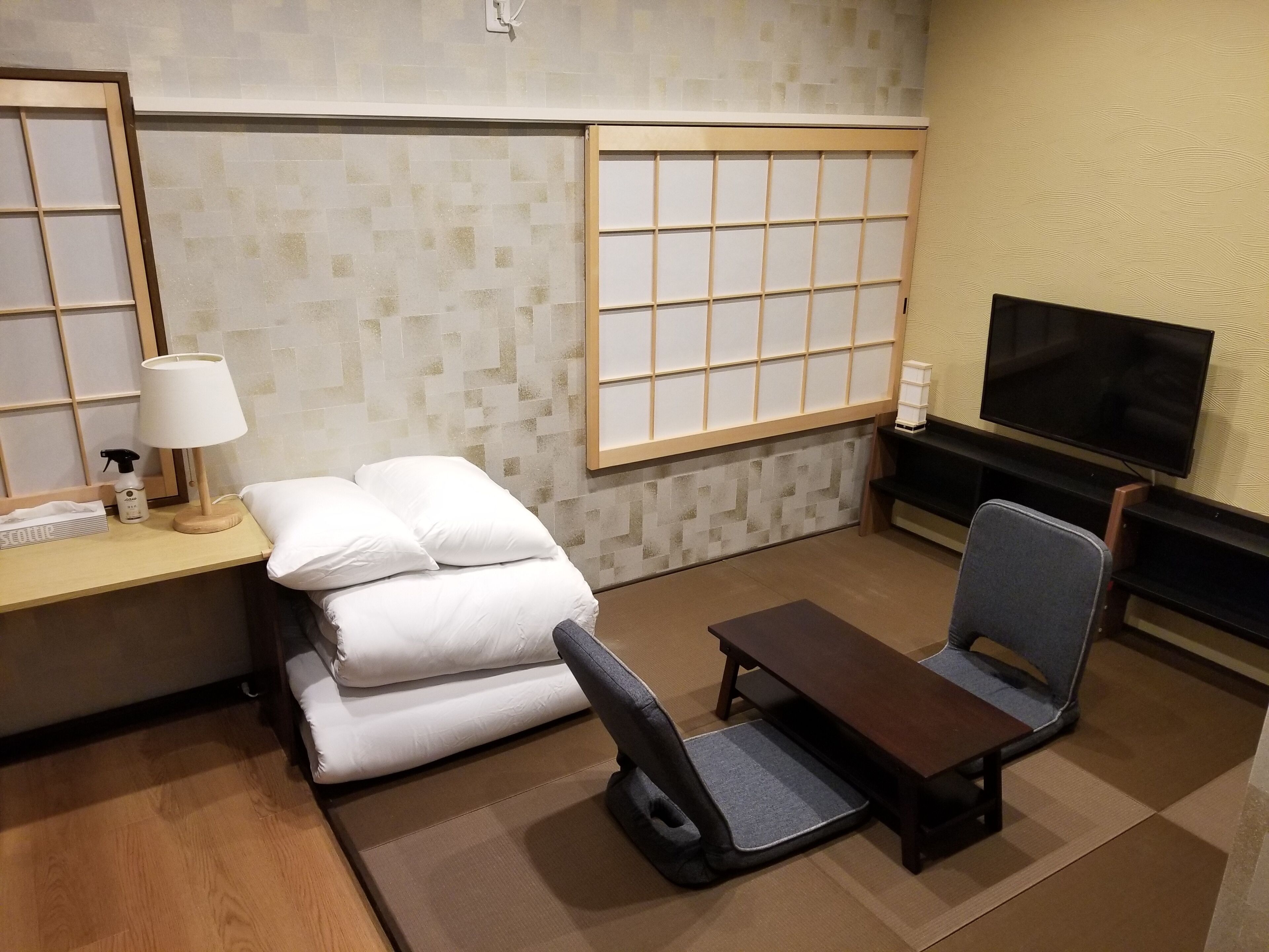A hotel room with a bed, a desk with a lamp, a TV, and a small table with a chair.
