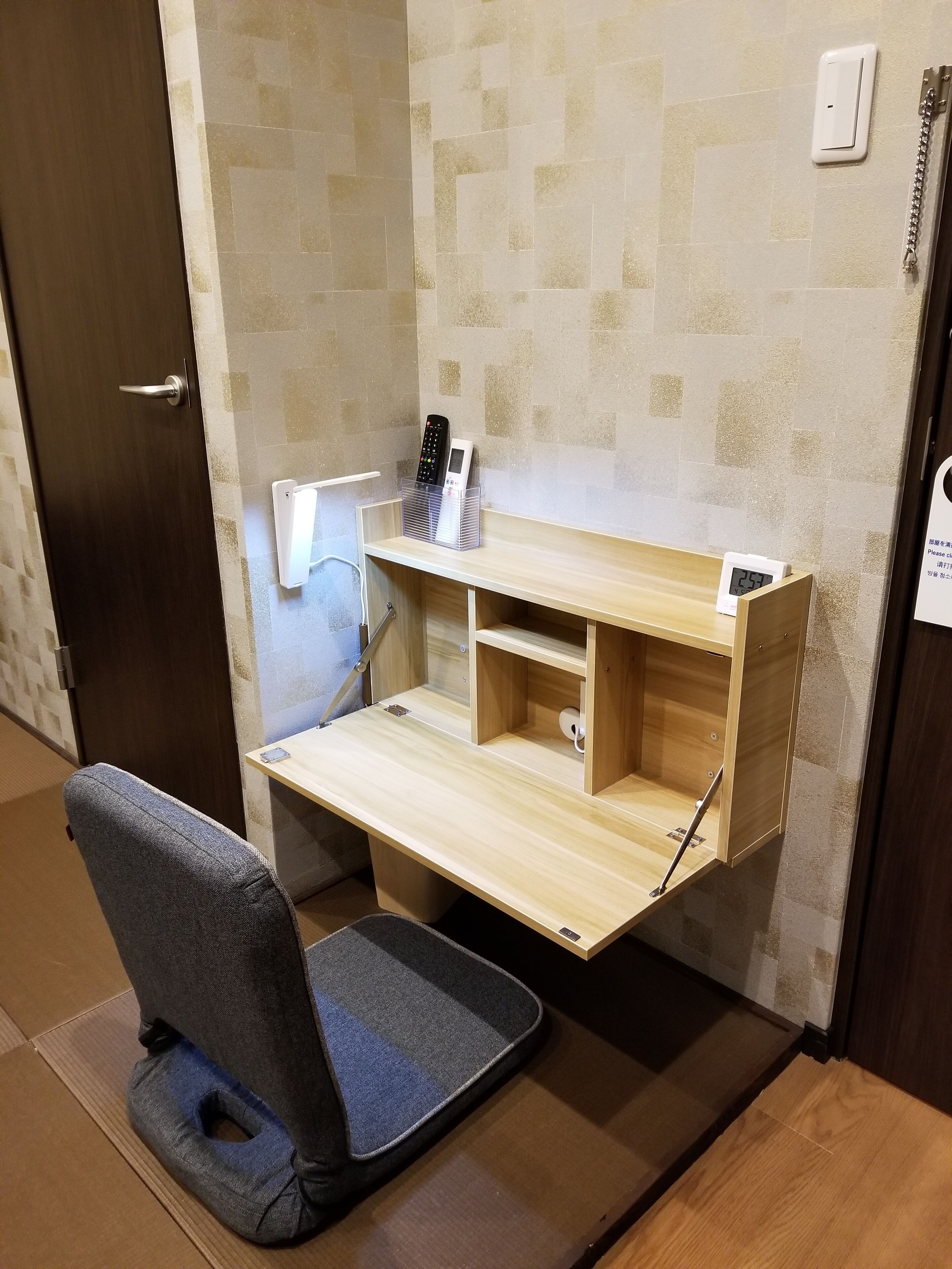 Une pièce de taille modeste, comprenant un bureau en bois, une chaise, une étagère fixée au mur avec un téléphone et une télécommande, et une horloge numérique.