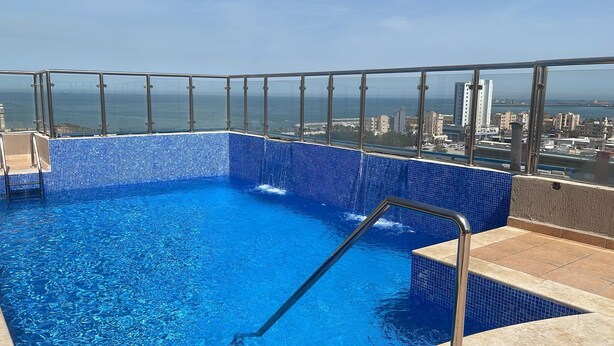 Ein Pool auf dem Dach mit klarem, blauem Wasser, umgeben von einem Metallgeländer, mit Blick auf eine Stadtlandschaft und den Ozean.