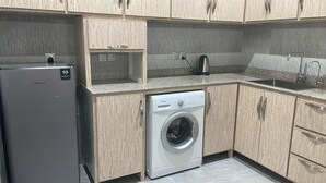 Eine kleine Küche mit einem Edelstahl-Kühlschrank, einer weißen Waschmaschine, Holzschränken, einer Granitarbeitsplatte und einem eingebauten Spülbecken.