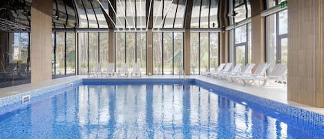 An indoor swimming pool with clear blue water, surrounded by lounge chairs and large windows offering a view of the outdoors.