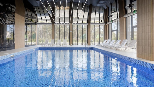 En inomhuspool med kristallklart blått vatten, omgiven av vita solstolar och stora fönster som ger utsikt över omgivningen.