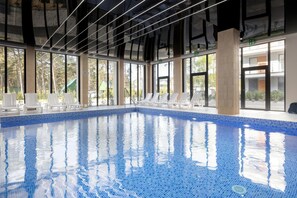 An indoor swimming pool with clear blue water, surrounded by large windows and white lounge chairs.