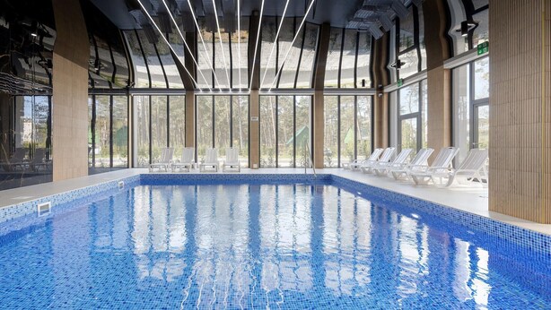 An indoor swimming pool with clear blue water, surrounded by white lounge chairs and large windows offering views of the outdoors. - Comfortable apartment, pool, close to the beach (Pobierowo)