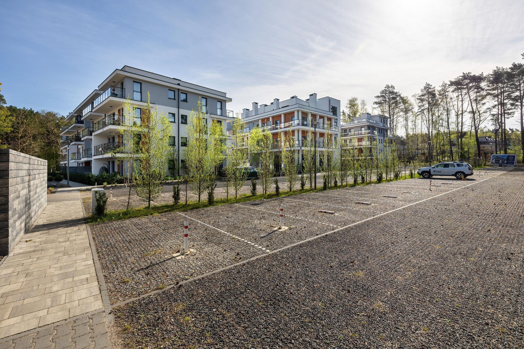 Ein modernes, mehrstöckiges Wohngebäude mit Balkonen, einem Kiesparkplatz und jungen Bäumen.