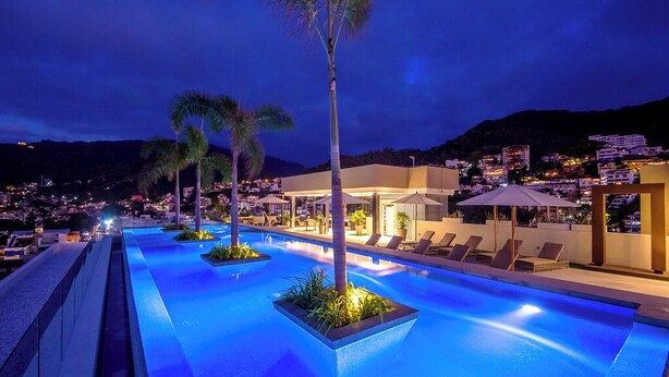 A nighttime swimming pool area with lounge chairs and palm trees, surrounded by buildings and a mountain in the background. - Casa Elizabeth Taylor | Rooftop Pool & Gym (Puerto Vallarta)