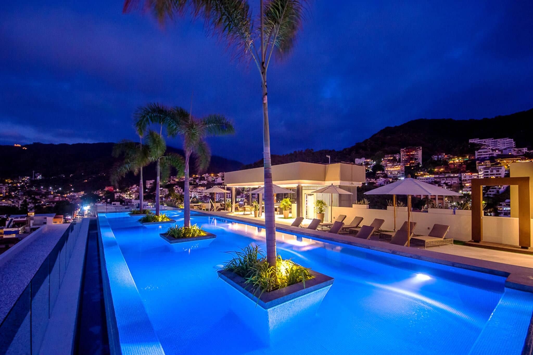 Zona de piscina nocturna con sillones de descanso y palmeras, rodeada de edificios y un telón de fondo montañoso.