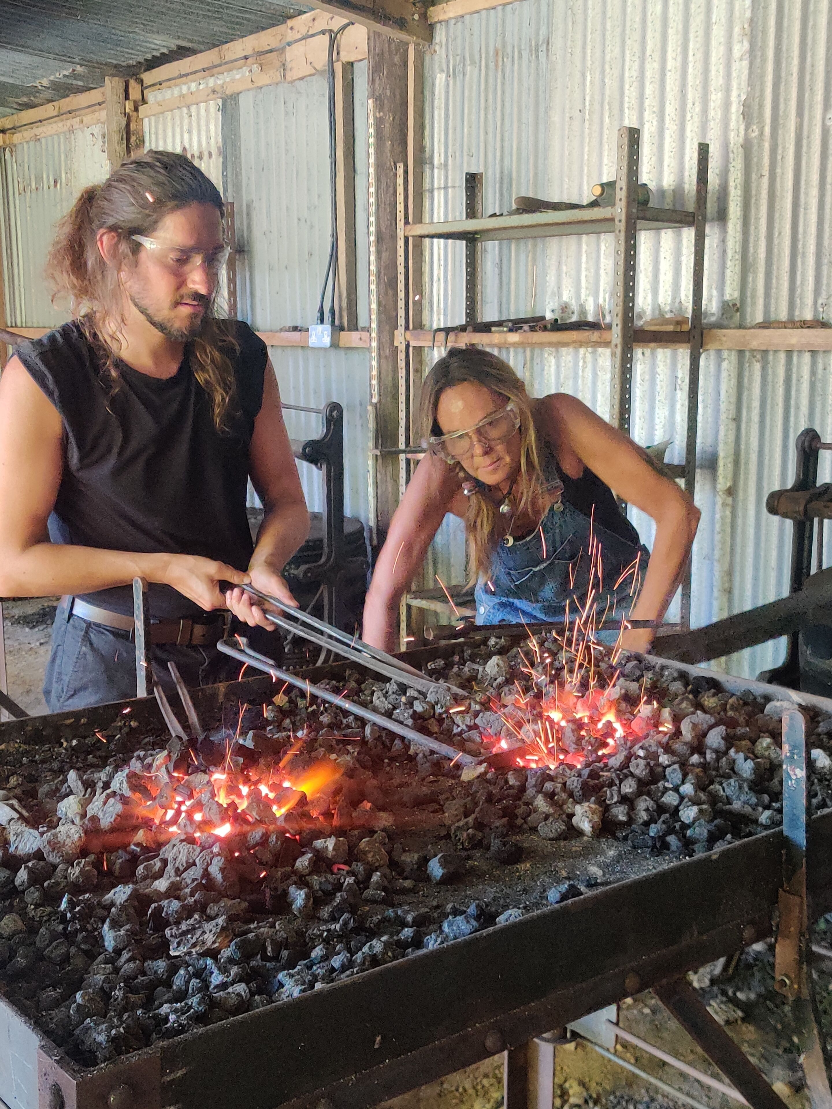 Two individuals working with metal tools and sparks at a forge.