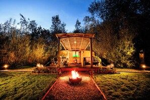 Um gazebo com uma lareira no centro, rodeado por um caminho de brita e vegetação.