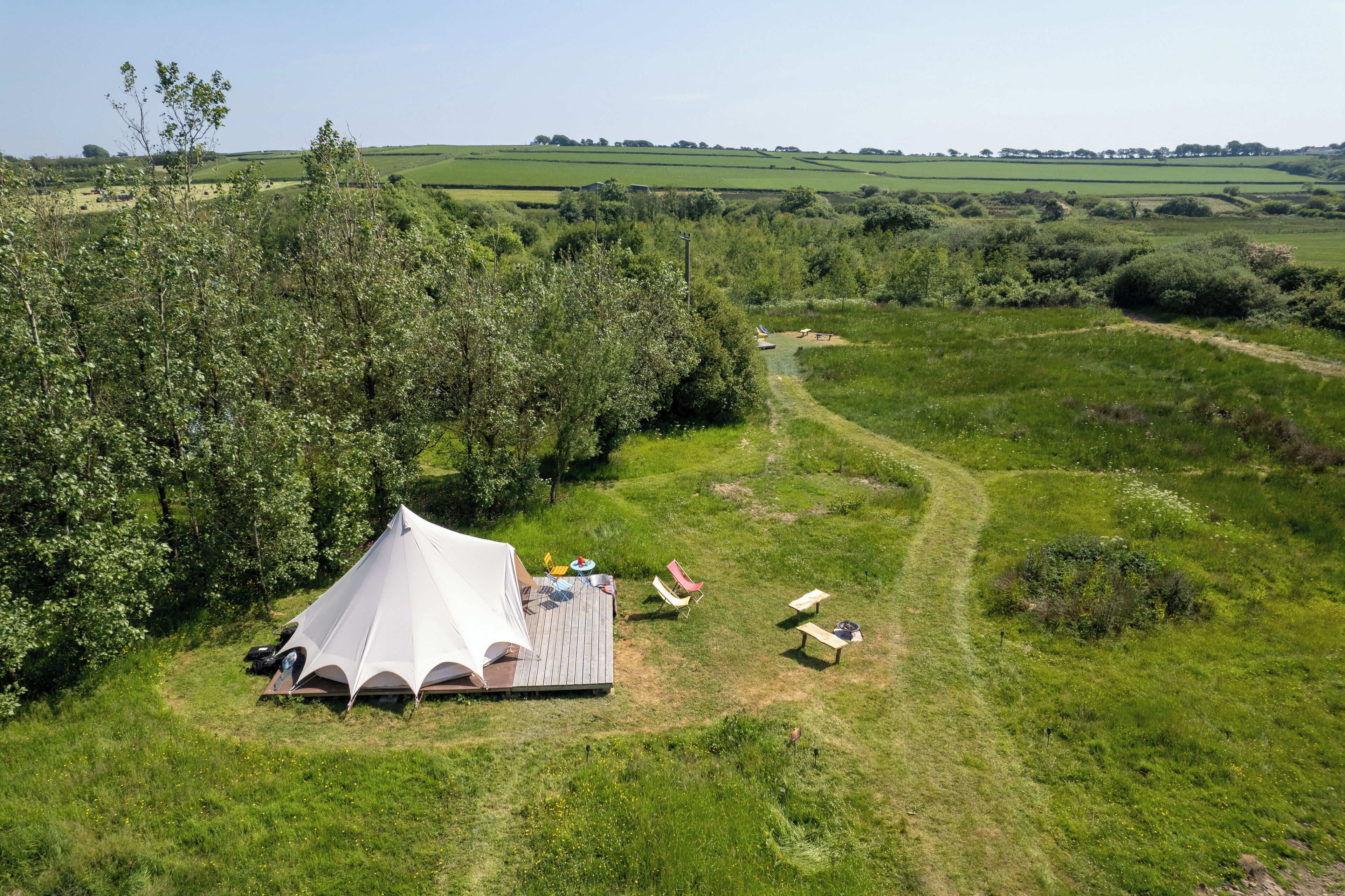 Een tentenveld met een picknicktafel, stoelen en een kleine boot in een grasveld met bomen en een pad.