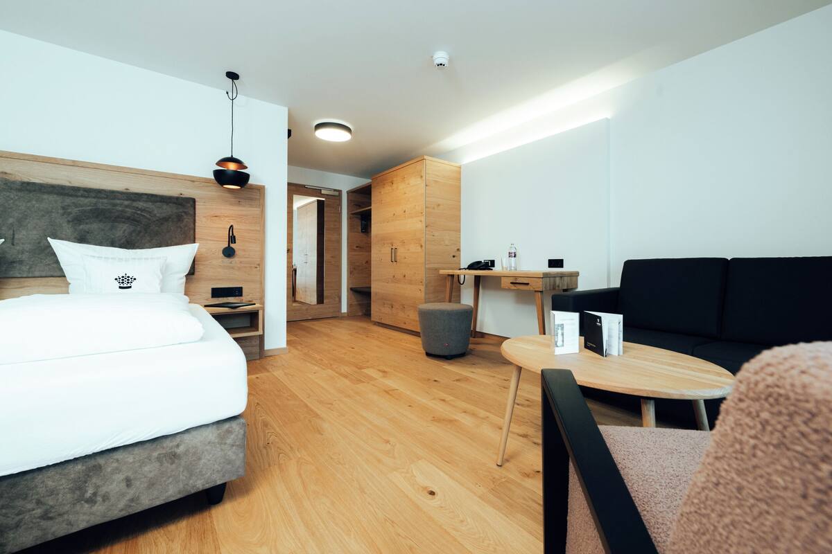 A modern hotel room with a bed, a wooden wardrobe, a small table, a black sofa, and a grey ottoman.