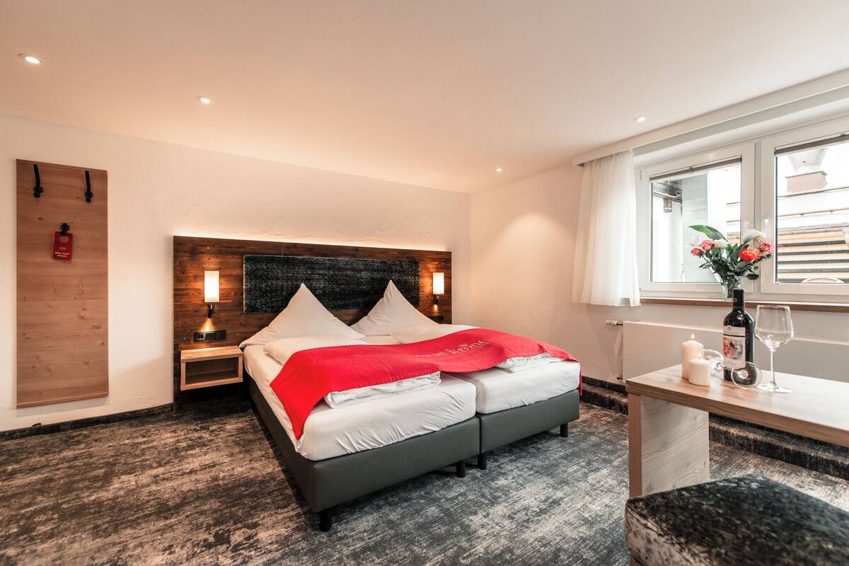 A modern hotel room with a large bed, bedside tables, a wooden headboard, a small desk with a vase of flowers, and a window with curtains.