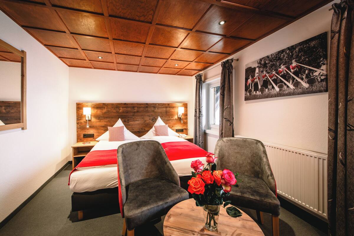A hotel room with a wooden ceiling, a bed with red and white bedding, two chairs, a small table with a vase of flowers, and a painting on the wall.