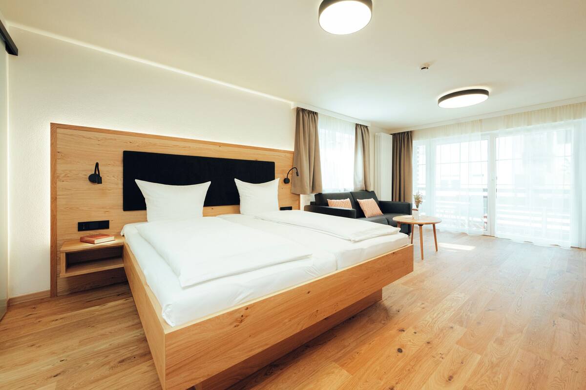 A modern hotel room with a large bed, wooden headboard, bedside tables, a small round table, and a sofa.