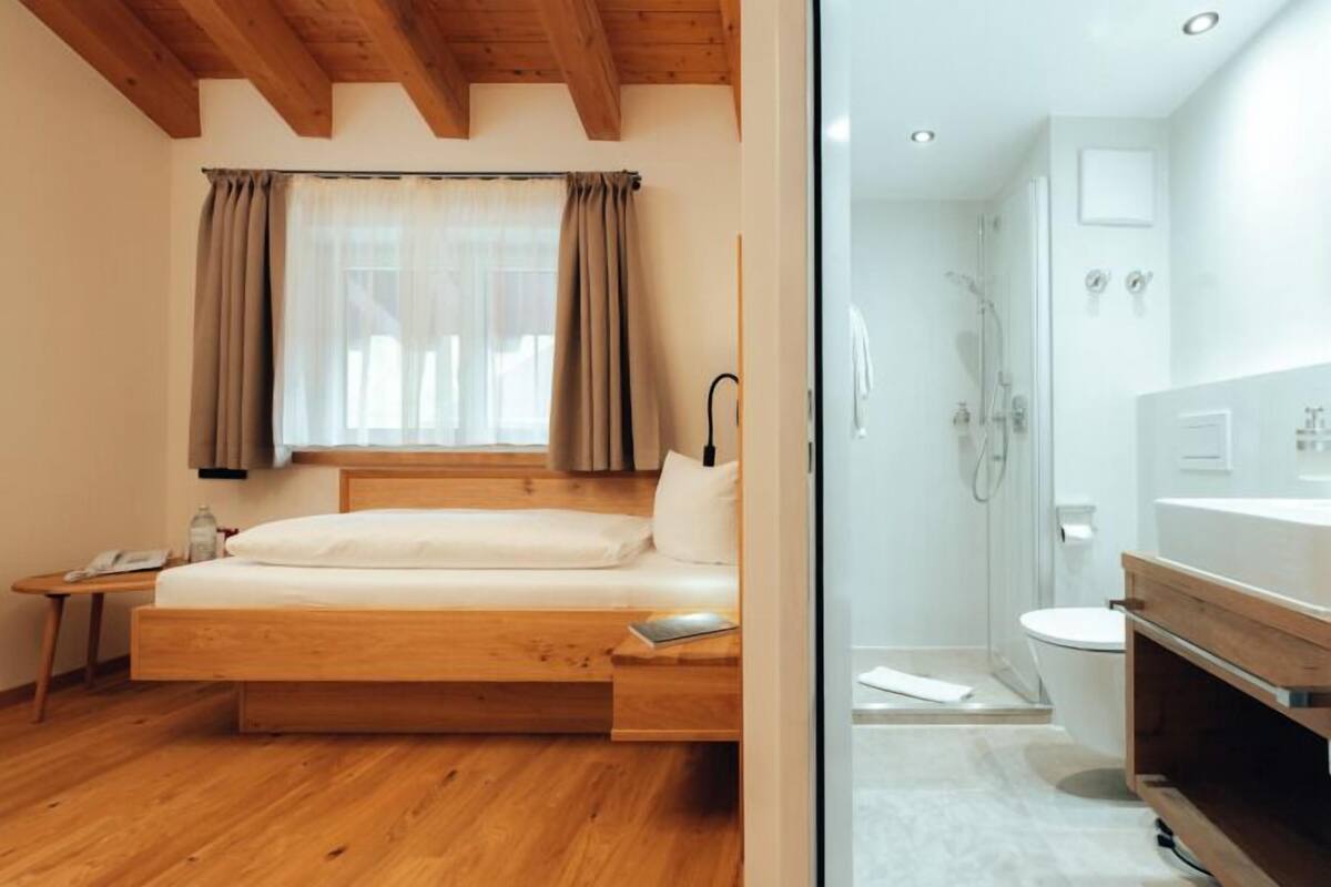 A bedroom with a wooden bed, a bedside table, a window with curtains, and a view of a bathroom with a shower and toilet.