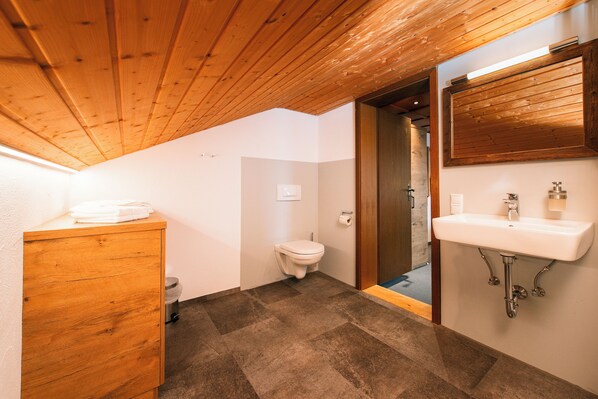 A bathroom with a wooden ceiling, a white toilet, a sink with a mirror, and a cabinet. - Hotel Tradizio (Mittelberg)