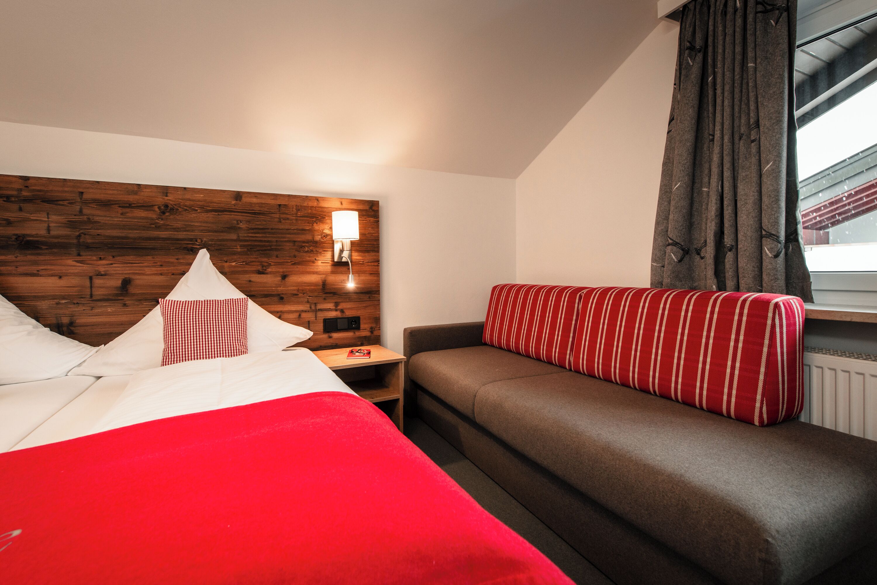 Una habitación de hotel con cabecera de madera, cama con ropa de cama roja, sofá gris con un cojín a rayas rojas y blancas, y una mesita de noche con lámpara.