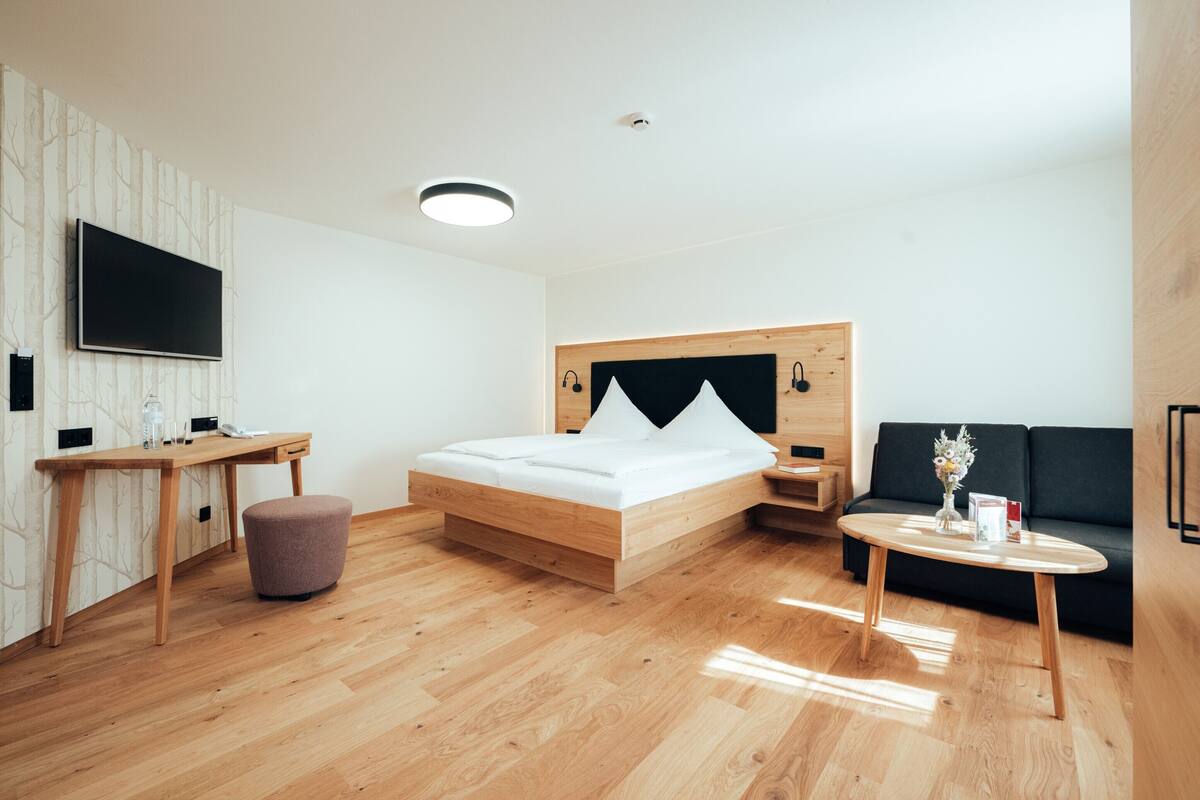 A modern hotel room with a wooden bed, a desk, a small ottoman, a sofa, and a TV mounted on the wall.