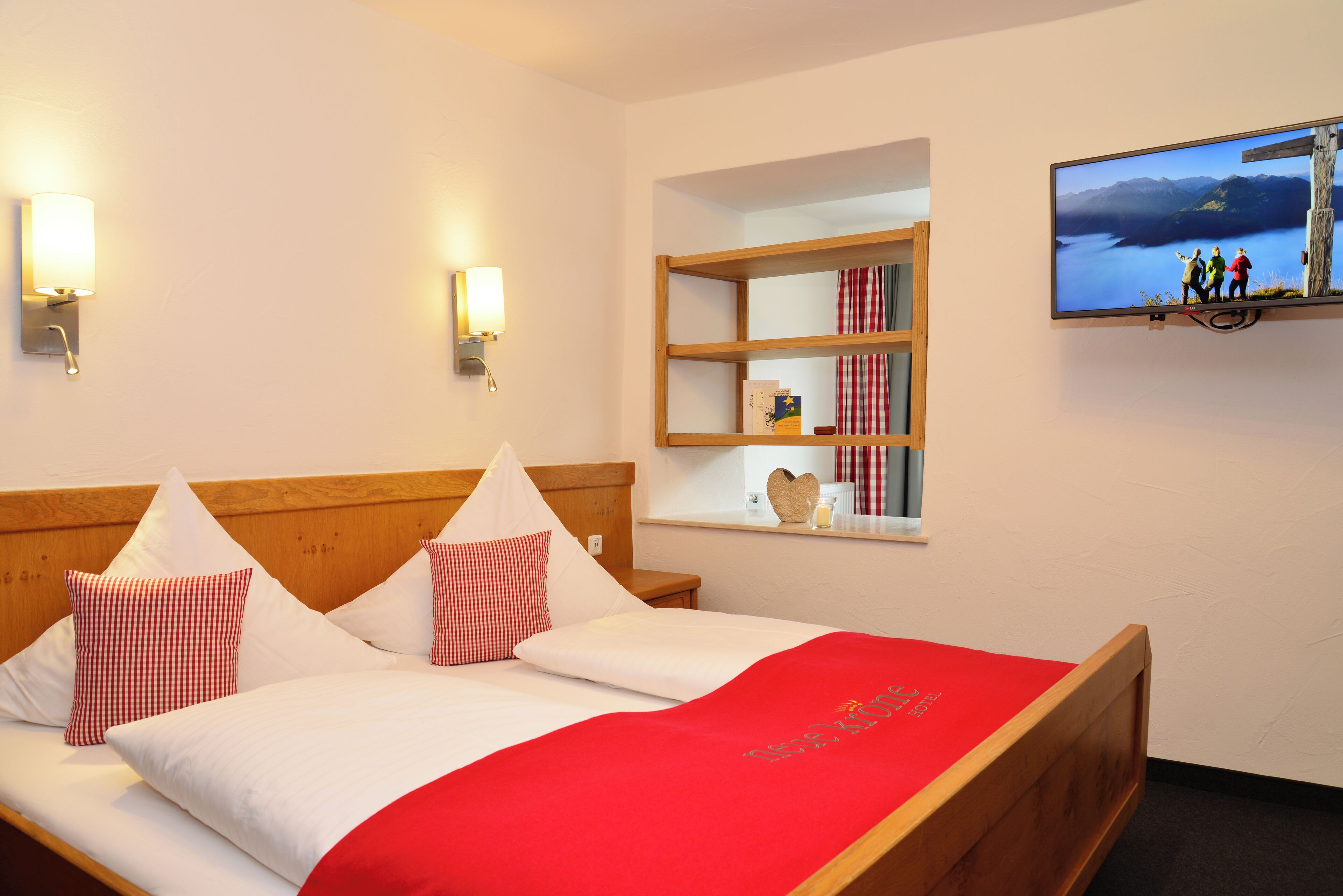 A hotel room with a wooden bed, white and red checkered pillows, a red bedspread, a wall-mounted TV, and a shelf with books and a heart-shaped decoration.