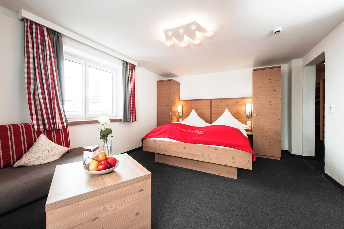 A hotel room with a wooden bed, a wooden nightstand, a bowl of fruit, a red blanket, and a window with curtains.