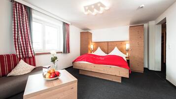 Una habitación de hotel con una cama de madera, una mesita de noche también de madera, un cuenco de fruta, una manta roja y una ventana con cortinas.