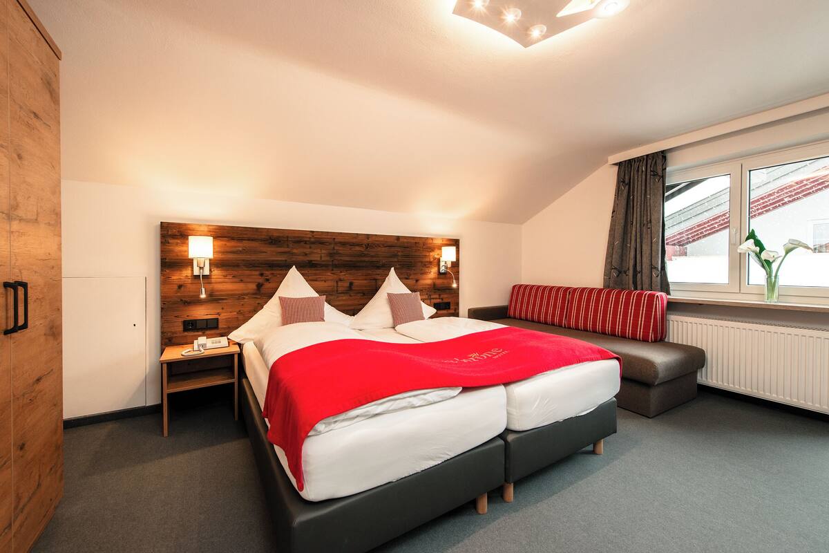 A modern hotel room with a wooden headboard, a large bed with a red blanket, a bedside table with a lamp, a sofa, and a window with curtains.