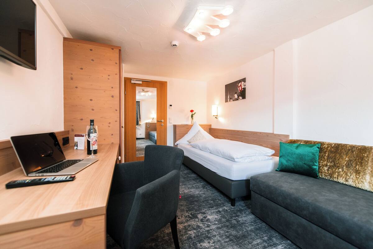 A hotel room with a wooden desk, a laptop, a bed, a sofa, and a small table with a wine bottle.