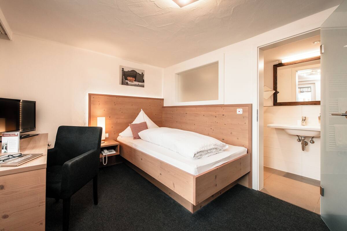 A hotel room with a wooden bed, a desk with a computer, a chair, a nightstand, a bedside table, a wall-mounted TV, a window, and a bathroom with a sink and mirror.