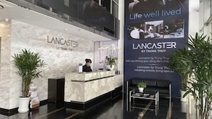 Un hall d’accueil d’hôtel avec un comptoir en marbre, un réceptionniste et une grande publicité pour « Lancaster by Trung Thuy », comprenant un slogan et des images d’un homme et d’une femme dans un espace de vie.