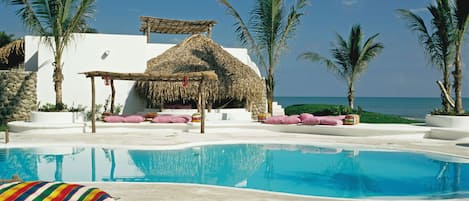A pool area with a thatched-roof structure, palm trees, and a clear view of the ocean.