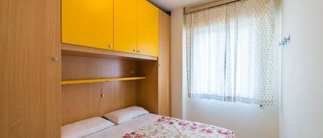 A bedroom with a bed, a yellow wardrobe, and a window with floral curtains.