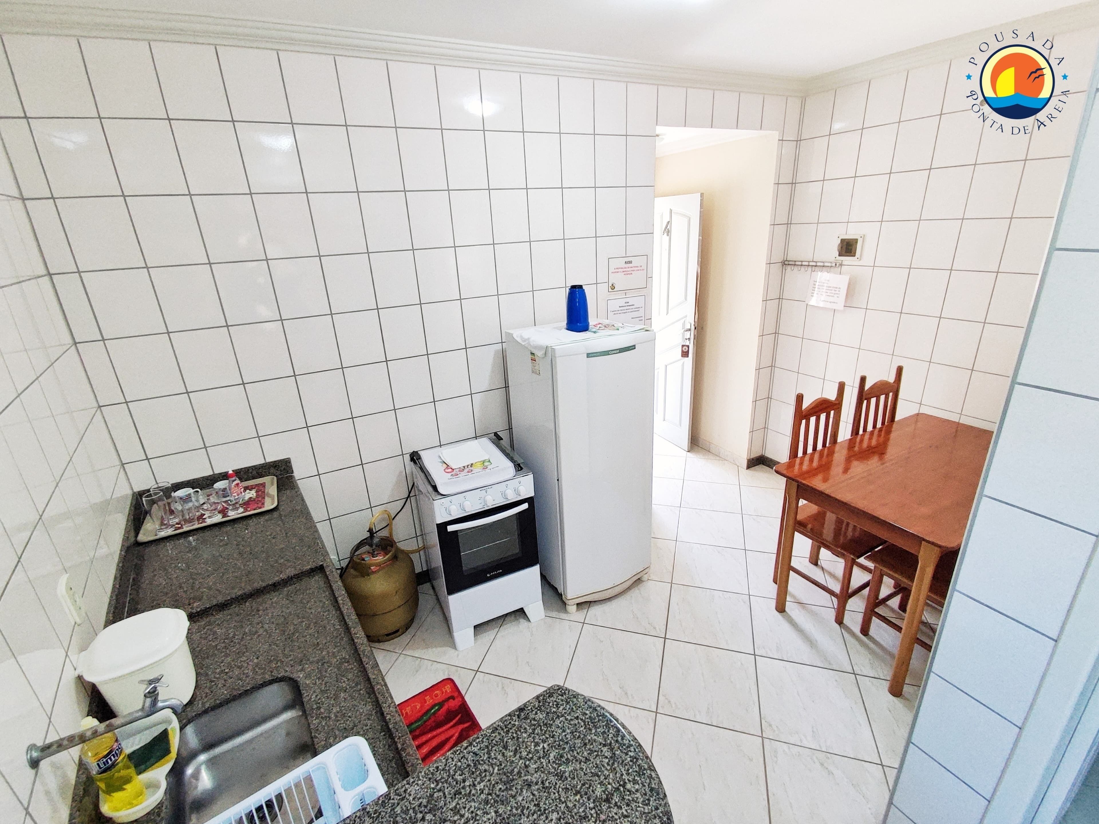 Cozinha compacta com paredes de azulejos brancos, fogão a gás, forno pequeno, geladeira, mesa de jantar com cadeiras e pia com torneira.